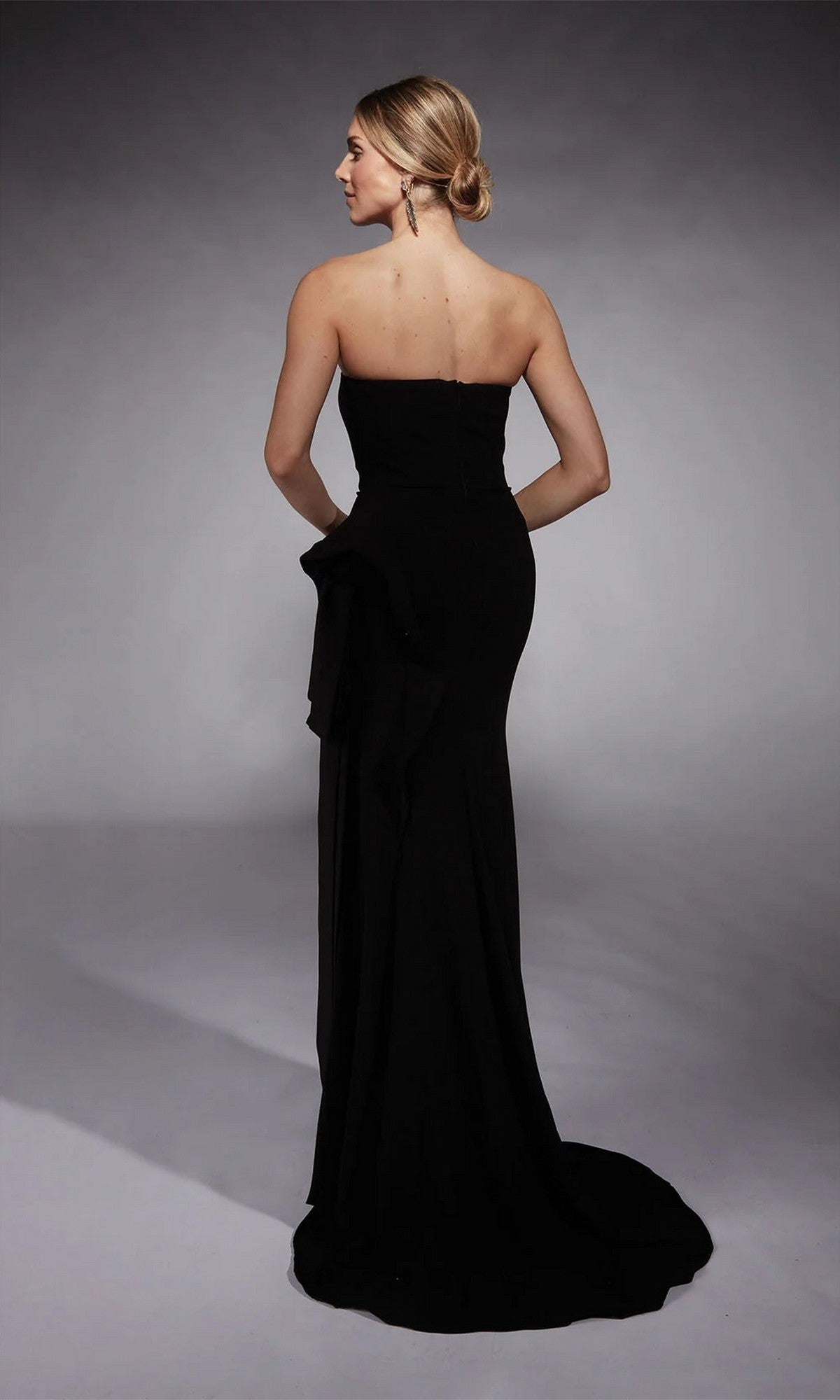 A woman wearing the Alyce 7114 long formal dress stands with her back to the camera against a neutral background, the elegant black strapless sheath gown flowing gracefully to the floor.